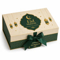 An elegant rectangular gift box for Eid Mubarak, featuring a cream-colored lid with gold patterns, hanging lantern illustrations, and a dark green central label. The front of the box is dark green with a gold pattern and a matching dark green satin ribbon tied in a bow, with a small hanging tag reading 'Eid Mubarak'.