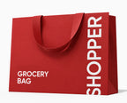 A red paper grocery shopping bag with two handles, featuring the word 'SHOPPER' printed vertically in white on the right side and 'GROCERY BAG' printed in white on the lower left, shown on a light gray background.